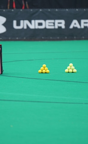 A sports field features a black and pink sports bag, two pyramids of yellow balls, and a portion of a person in dark athletic gear. The background displays a banner with the Under Armour logo.