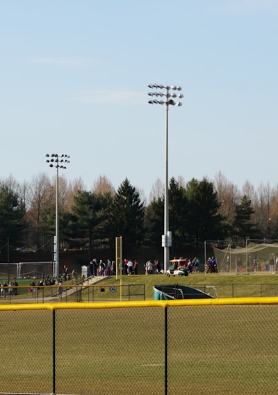 A sports field with a group of people engaging in athletic activities in the background. There is sports equipment like a soccer goal and a scoreboard visible. A chain-link fence runs across the foreground, and a statue or sculpture resembling an eagle is also seen. Trees and tall floodlights are positioned behind the field.
