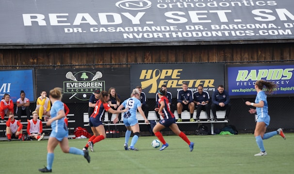 Players engage in a competitive soccer game on a field while others are seated on the bench watching attentively. The action is taking place in front of a wooden structure displaying multiple advertisements, including orthodontics, lacrosse, and field hockey.