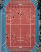 An aerial view of a large, vibrant sports complex featuring a red basketball court with multiple hoops and a surrounding blue running track. Several people are scattered across the area, engaged in various activities.
