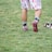 A person wearing sports shoes and patterned shorts stands on a grassy field, with various sports equipment like gloves and a helmet placed on the ground nearby.