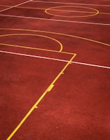 A sports court with a reddish-brown surface and bright yellow and white lines. The lines form geometric shapes, including rectangles and circles, suggesting markings for games like basketball or netball.