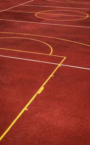 A sports court with a reddish-brown surface and bright yellow and white lines. The lines form geometric shapes, including rectangles and circles, suggesting markings for games like basketball or netball.