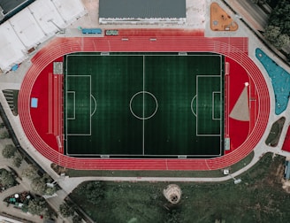 An aerial view of a large sports complex featuring a central green football field surrounded by a red running track. Adjacent to the track, there is a parking area, buildings, and pathways. Nearby, there are additional sports courts in different colors, including a blue swimming pool and orange basketball courts.