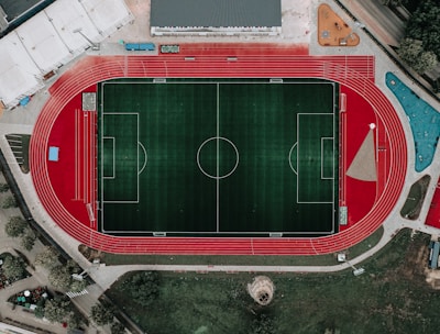 An aerial view of a large sports complex featuring a central green football field surrounded by a red running track. Adjacent to the track, there is a parking area, buildings, and pathways. Nearby, there are additional sports courts in different colors, including a blue swimming pool and orange basketball courts.