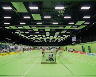 A sports hall with bright green flooring and ceiling panels. There is a game in progress with players moving quickly, suggesting a fast-paced sport like floorball or indoor hockey. Advertising banners line the walls, and a scoreboard is visible in the background.
