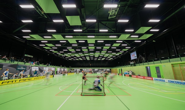 A sports hall with bright green flooring and ceiling panels. There is a game in progress with players moving quickly, suggesting a fast-paced sport like floorball or indoor hockey. Advertising banners line the walls, and a scoreboard is visible in the background.