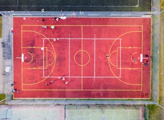 An aerial view of a sports court with a vibrant red surface and yellow lines marking different sections for basketball and soccer. Several people are present on the court, some engaged in activities, while others are standing along the perimeter. The court is enclosed by a metal fence and surrounded by grass on all sides.