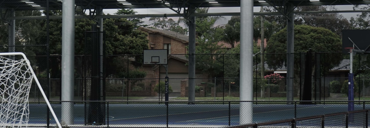 An outdoor sports facility featuring a running track and a soccer goal. The track is red, surrounded by a black metal fence, and the field is covered with artificial green turf. In the background, a basketball court is visible under a covered roof with metal supports. The sky is overcast, adding a moody atmosphere.