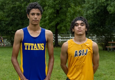 Two young athletes wearing sleeveless sports tops stand on a grassy field. The athlete on the left is wearing a blue top with the word 'TITANS' while the one on the right wears a yellow top with 'ASIJ'. The background features green trees and a white fence, with a bench visible on the right.