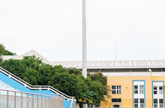 A sports stadium with a red running track, blue seating area, and a large floodlight. The background features a yellow building with blue and white accents, surrounded by green trees. The atmosphere is clean and empty, with clear skies above.