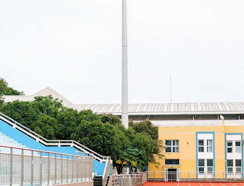 A sports stadium with a red running track, blue seating area, and a large floodlight. The background features a yellow building with blue and white accents, surrounded by green trees. The atmosphere is clean and empty, with clear skies above.