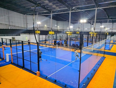 An indoor sports facility featuring several padel courts with blue playing surfaces and orange sidelines. There are players engaged in a game on one of the courts, while the others appear empty. The area is well-lit with overhead lighting and enclosed by a high ceiling and metal structure.