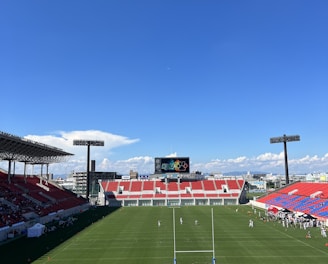 A large sports stadium with red and blue seating overlooks a green field with a goalpost at the center. A few people are scattered across the field, with some tents and a gathering of individuals on the right side. The sky is clear and blue with some clouds visible in the distance. A large screen or scoreboard is situated at the far end of the field.
